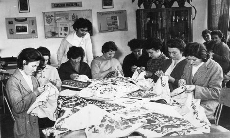 Fekete-fehér archív fotó: asztal körül ülő nők hímeznek nagyméretű, mintás textileken, középen álló szakkörvezető nő irányít. Háttérben falon kiállított hímzések, fotók.