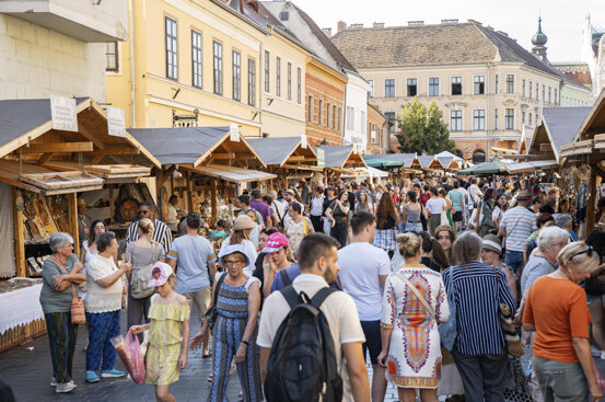 Színes fotó: nyári fesztiváli forgatag a Budai Várnegyedben. Két sorban felállított fa árusítóstandok között sűrű tömeg sétál, jobbra a Mátyás-templom tornya látszik. A Mesterségek Ünnepe rendezvénye.